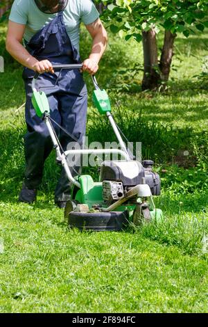 Un giardiniere falcia erba verde con un falciatore a benzina industriale, tende un prato verde tra gli alberi in un giardino di primavera. Immagine verticale, spazio di copia. Foto Stock