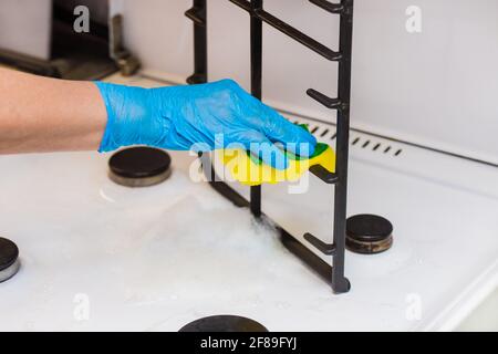 Mano di una donna in un guanto domestico in lattice spugne la griglia di una stufa a gas in cucina, primo piano. Foto Stock