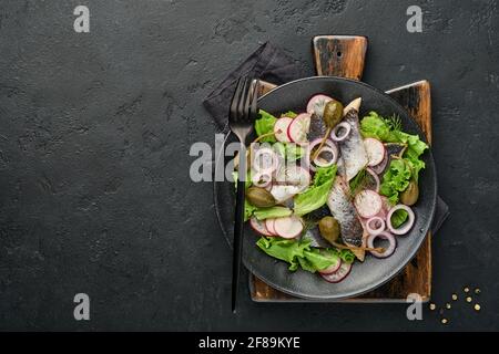 Aringhe salate con spezie, capperi, erbe e insalata di cipolla su piatto nero su sfondo scuro con spazio copia. Pesce marinato a fette. Cibo con u sano Foto Stock