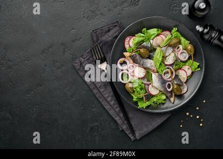 Aringhe salate con spezie, capperi, erbe e insalata di cipolla su piatto nero su sfondo scuro con spazio copia. Pesce marinato a fette. Cibo con u sano Foto Stock