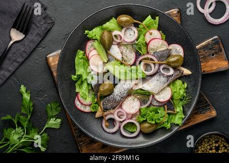 Aringhe salate con spezie, capperi, erbe e insalata di cipolla su piatto nero su sfondo scuro con spazio copia. Pesce marinato a fette. Cibo con u sano Foto Stock