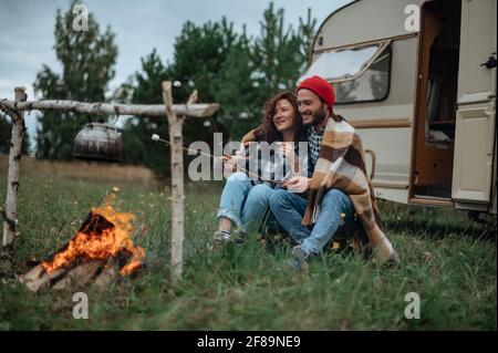 Accoppia in un plaid a scacchi arrostendo marshmallows sul fuoco vicino alla casa del rimorchio. Foto Stock