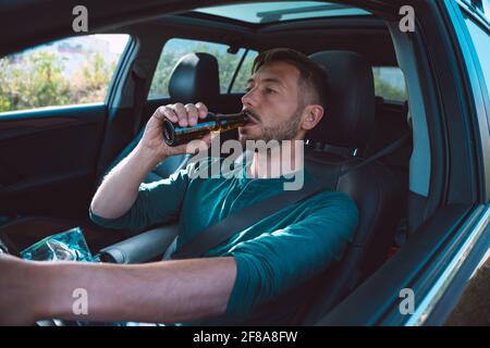 Driver ubriaco. Giovane uomo che beve birra durante la guida di un'auto. Conducente sotto l'influenza dell'alcol. Concetto di guida pericolosa. Foto Stock