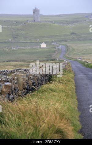 Muro di pietra e strada che conduce a Mist e rovine del castello in distanza, Irlanda rurale Foto Stock