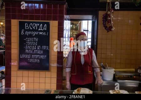 Mosca, Russia. 11 aprile 2021. Una donna di vendita indossa una maschera al negozio Yeliseyevsky il giorno della sua chiusura. Considerato il più antico negozio di alimentari di Mosca, fu fondato dal mercante Grigory Yeliseyev nel 1901 come Negozio di Elisev e Cantine di vini russi e stranieri. Il 5 febbraio di quest'anno, il negozio ha segnato 120 anni dalla sua apertura. Il banner recita 'sempre in vendita insalate, sottaceti e piatti a base di carne, pesce, pollame' Foto Stock