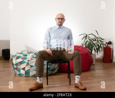 Giovane uomo di affari alla moda calvo si siede sulla sedia con mani su gambe larghe in camera con pareti luminose e divani pigri vaso fiore fuoco extin Foto Stock