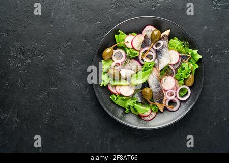 Aringhe salate con spezie, capperi, erbe e insalata di cipolla su piatto nero su sfondo scuro con spazio copia. Pesce marinato a fette. Cibo con u sano Foto Stock