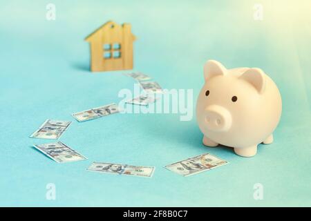 Il concetto di risparmio di denaro per acquistare un appartamento, una casa o un'altra proprietà residenziale. Piggy bank e casa di legno su sfondo blu. Foto Stock