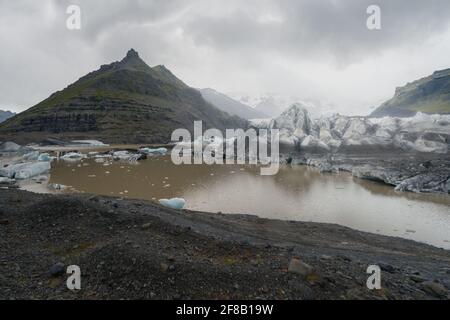 Ghiaccio che galleggia nelle acque fangose del lago glaciale con ghiacciaio sullo sfondo in una giornata nuvolosa e piovosa. Fjallsarlon, Islanda. Foto Stock