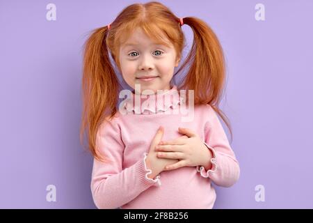 Bambina che tiene le mani sul torace, sentendo gratitudine o sognando e guardando la macchina fotografica, isolato su sfondo viola. Copia spazio Foto Stock