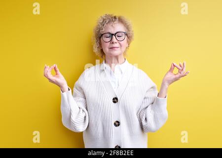 Calma sicura donna anziana in occhiali meditando isolato su sfondo giallo studio, in piedi con gli occhi chiusi, concentrato sulla mente. Foto Stock