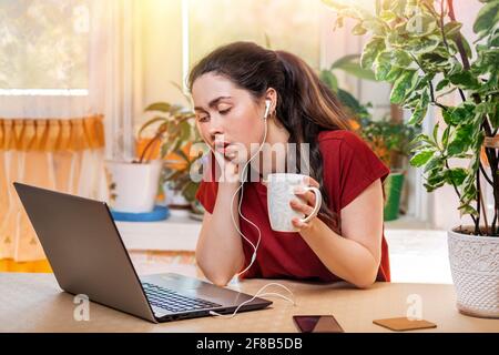 Auto-isolation.UNA giovane bella donna caucasica, con uno sguardo annoiato, tiene una tazza nelle sue mani, e guarda sonnolentemente il suo laptop, con le cuffie in lei Foto Stock