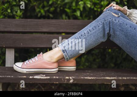 Primo piano delle gambe di una donna caucasica che indossa jeans e le sneaker che si stendono su una panca Foto Stock
