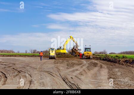 Il progetto è in corso, l'escavatore sta riempiendo un dumper di terreno e il rullo stradale si sta appiattendo, livellando, equivale al cantiere. Foto Stock