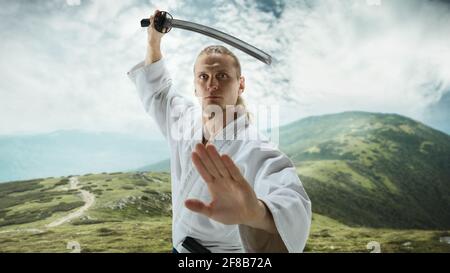 Giovane uomo, insegnante che combatte Aikido, allenando arti marziali sul prato in montagna di fronte Foto Stock