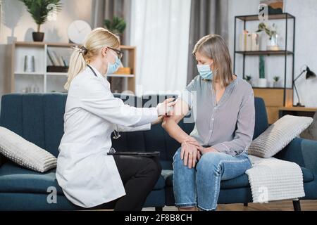 Professionista sicuro medico femminile in maschera facendo l'iniezione di vaccino o di insulina alla donna anziana paziente in maschera, visitandola a casa. Sanità, vaccinazione, pandemia di covid-19. Foto Stock