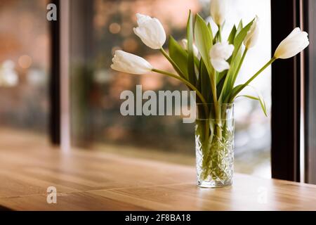 Vaso con fiori bianchi su grande sfondo finestre con tavolo in legno. Concetto di intimità domestica. Bouquet di tulipani in vaso di vetro Foto Stock