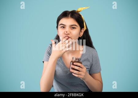 Adolescente Indial ragazza mangiare crema di cioccolato dal vaso, sentirsi soddisfatto, come il suo dito su sfondo blu studio Foto Stock