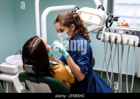 La giovane donna si siede nella sedia del dentista e il dentista esamina attentamente i denti del paziente. Il dentista consulta un paziente seduto su una sedia a t. Foto Stock