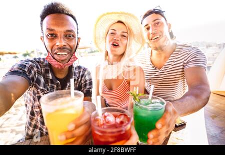 Trio di persone multirazziali che prendono selfie con maschere aperte al viso beach bar - nuovo concetto di vita normale con ubriaco amici che si divertono insieme Foto Stock