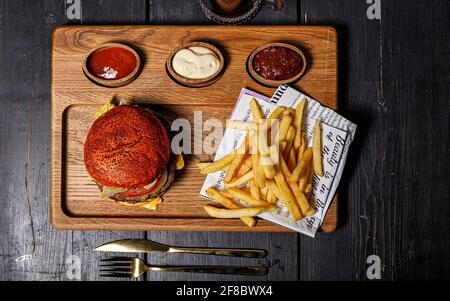 Delizioso hamburger doppio di manzo e un servizio di patatine fritte su un tagliere di legno con tre tipi di salsa. Sfondo rustico scuro. Fast food Foto Stock