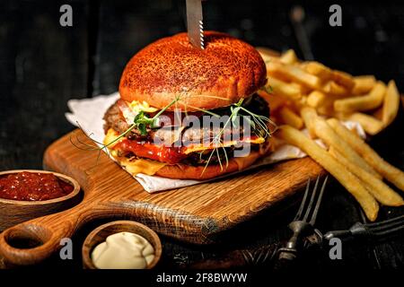 Appetitoso hamburger di manzo doppio e una porzione di patatine fritte su un tagliere di legno. Rustico sfondo scuro. Fast food fotografia di natura morta. Primo piano Foto Stock