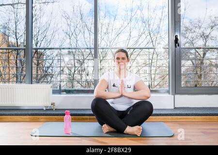 Bella donna più dimensioni sedersi nella palestra leggera sport sala in lotus e namaste yoga posa a piedi nudi con bottiglia d'acqua Foto Stock