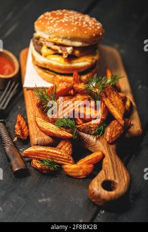 Appetitoso hamburger di manzo doppio e una porzione di patatine fritte su un tagliere di legno. Rustico sfondo scuro. Fast food fotografia di natura morta. Sho. Verticale Foto Stock