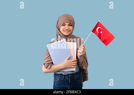 Teen girl in hijab musulmano tradizionale tenendo bandiera turca e libri di testo su sfondo blu. Istruzione per le donne arabe Foto Stock