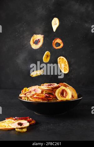 Prugne secche, kiwi, pesca, patatine di frutta arancione che volano in ciotola nera su sfondo nero, asciugando a casa. Snack vegan senza zucchero cibo. Foto Stock