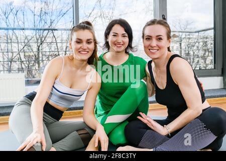 Tre donne di sport sorridenti che si siedono sui tappetini a piedi nudi in studio di fitness leggero, esercizi di allenamento di gruppo Foto Stock