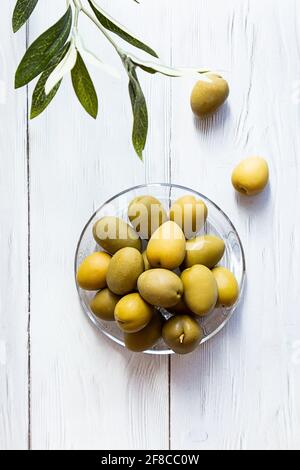 Una ciotola di olive verdi. Minimo fermo di una ramificazione di olive e di una coppa di olive su un tavolo di legno bianco. Spazio di copia Foto Stock