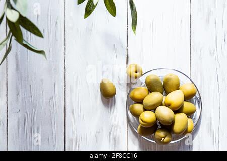 Una ciotola di olive verdi. Minimo fermo di una ramificazione di olive e di una coppa di olive su un tavolo di legno bianco. Spazio di copia Foto Stock