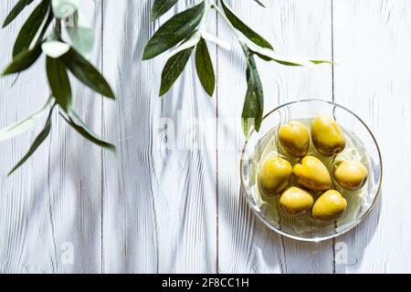 Una ciotola di olive verdi sottaceto. Minimo fermo di una ramificazione di olive e di una coppa di olive su un tavolo di legno bianco. Spazio di copia Foto Stock