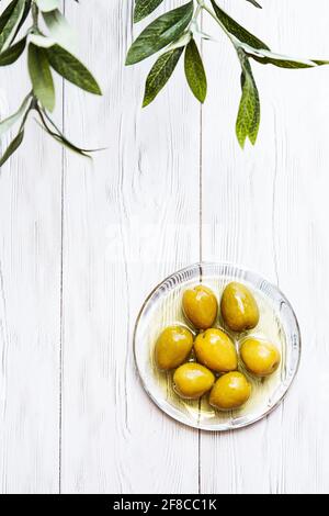 Una ciotola di olive verdi sottaceto. Minimo fermo di una ramificazione di olive e di una coppa di olive su un tavolo di legno bianco. Spazio di copia Foto Stock