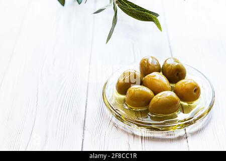 Una ciotola di olive verdi sottaceto. Minimo fermo di una ramificazione di olive e di una coppa di olive su un tavolo di legno bianco. Spazio di copia Foto Stock