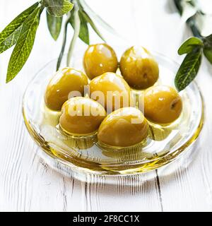 Una ciotola di olive verdi sottaceto. Minimo fermo di una ramificazione di olive e di una coppa di olive su un tavolo di legno bianco. Spazio di copia Foto Stock