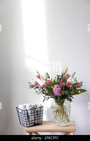 Un bouquet minimalista di tulipani bianchi, eustoma rosa, giacinto, eucalipto in vaso di vetro scanalato su una sedia. Piccoli asciugamani bianchi per le mani e il viso Foto Stock