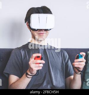 Giocare a casa con un visore VR, realtà virtuale, adolescente / giovane uomo negli anni '20, ambiente in salotto, seduto su un divano Foto Stock