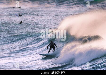 Un surfista che cavalca una grande onda come spray è soffiato fuori dalla cresta dal forte vento offshore in Fistral Bay a Newquay in Cornovaglia. Foto Stock