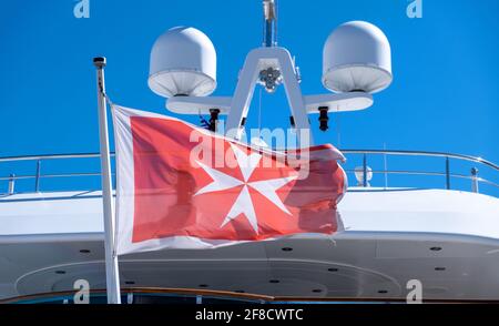 Vecchia bandiera maltese croce bianca su sfondo rosso sventolando su poppa yacht. Barca di lusso ancorata al porto turistico in Grecia. Sfondo blu cielo, vista ravvicinata. Foto Stock