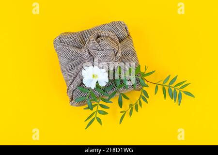 Regali Furoshiki. Eco-friendly avvolti in tessuto di lino con fiori freschi e foglie su sfondo giallo alla moda. Spazio di copia. Disposizione piatta. Foto Stock