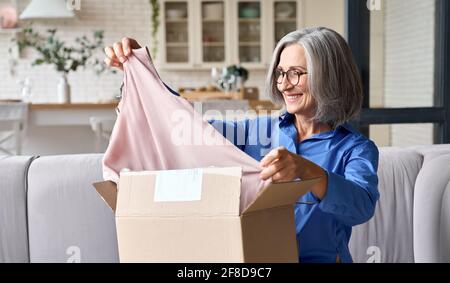 Senior metà anni 60 donna apertura scatola di pacchi a casa sul divano. Foto Stock