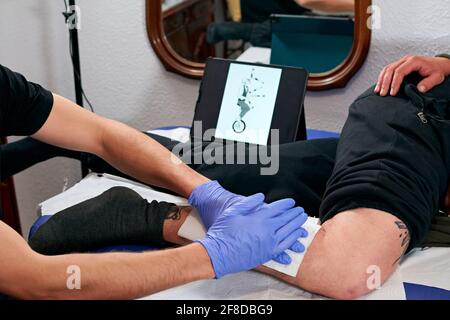 Mani di un artista tatuaggio in guanti blu che puliscono il tatuaggio appena completato sulla gamba di un uomo Foto Stock