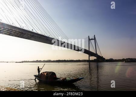 Kolkata, Bengala Occidentale, India - Gennaio 2018: Silhouette di un uomo che naviga in una barca di legno sotto il ponte Howrah iconico sopra il fiume Hooghly nel cit Foto Stock