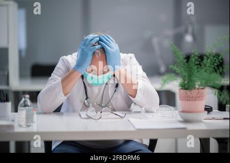 Durante la pandemia, un medico di sesso maschile anziano è molto stanco dal lavoro, si siede alla sua scrivania nell'ufficio del residente e tiene la testa. Foto Stock