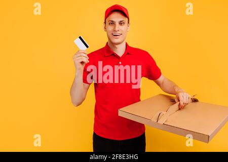 Servizio di consegna uomo in T-shirt rossa e cappuccio, dà ordine di cibo e tiene la scatola della pizza e carta di credito su sfondo giallo isolato Foto Stock