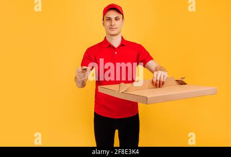 Servizio di consegna uomo in T-shirt rossa e cappuccio, dà ordine di cibo e tiene la scatola della pizza e carta di credito su sfondo giallo isolato Foto Stock