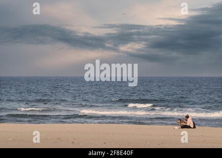 Donna seduta con il cane in riva al mare sulla spiaggia di Sagunto con il molo di Port De Sagunt sullo sfondo, Valencia, Spagna Foto Stock