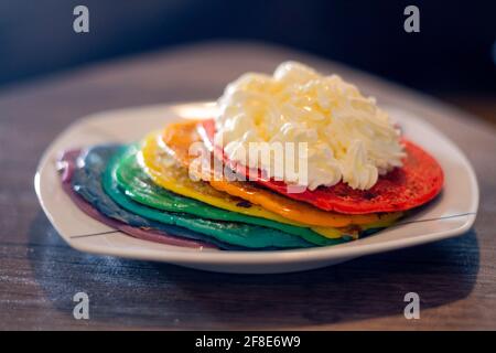 Primo piano di un piatto con frittelle color arcobaleno con un dollop di panna montata su un tavolo di legno Foto Stock
